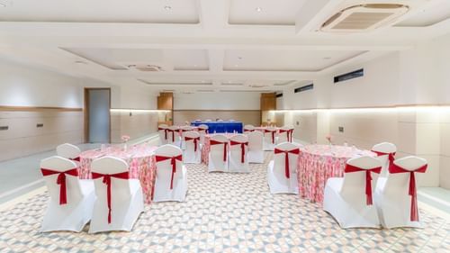 Banquet Hall at Pride Elite Dwarka featuring chairs and table decorated with ribbons and table cloth respectively. There is also a conference table with chairs at one end.
