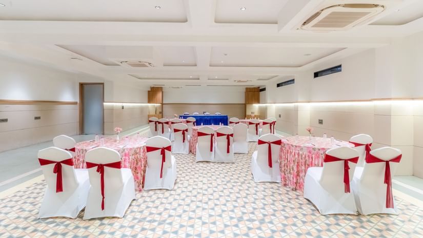 Banquet Hall at Pride Comfort Dwarka featuring chairs and table decorated with ribbons and table cloth respectively. There is also a conference table with chairs at one end.