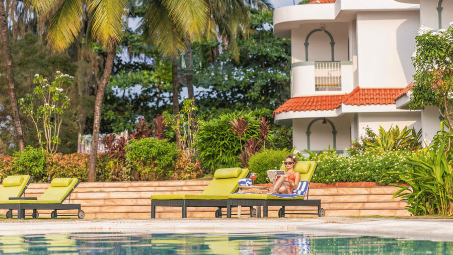 Guests relaxing on sun loungers beside a pool amidst tropical greenery | South Goa Hotel near Beach