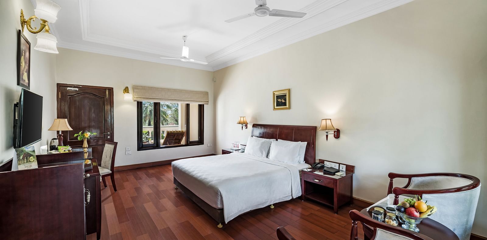 An image of the Garden room with river view featuring a queen size bed, bed side tables, lamps, ceiling fan, flat screen TV, and chairs - GReaT Trails River View Resort Thanjavur By GRT Hotels