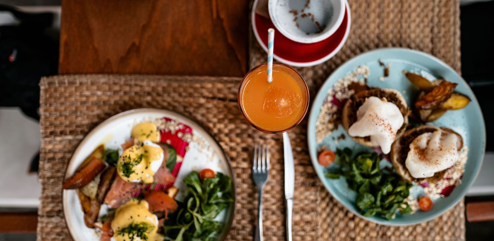 Overhead view of a hearty breakfast or brunch spread, featuring eggs Benedict, salad, a savoury main course on a blue plate, orange juice, and a cup of tea or coffee, all set on a woven placemat.