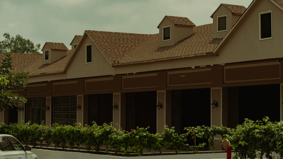 A tan building features multiple small dormer windows on a brown tiled roof, with a row of green potted plants lining the walkway below at Jivana Wellness Retreat, Bhopal.