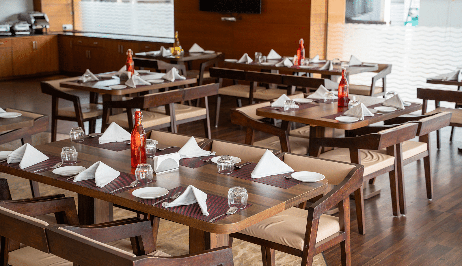 An elegant indoor restaurant setup with polished wooden tables, folded napkins, and large windows providing natural light - Hotel Udai Median, Udaipur.