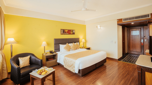 A modern bedroom with a neatly-made bed, yellow accent wall, leather armchair, coffee table with a fruit plate, and wooden flooring at The Piccadilly Lucknow.