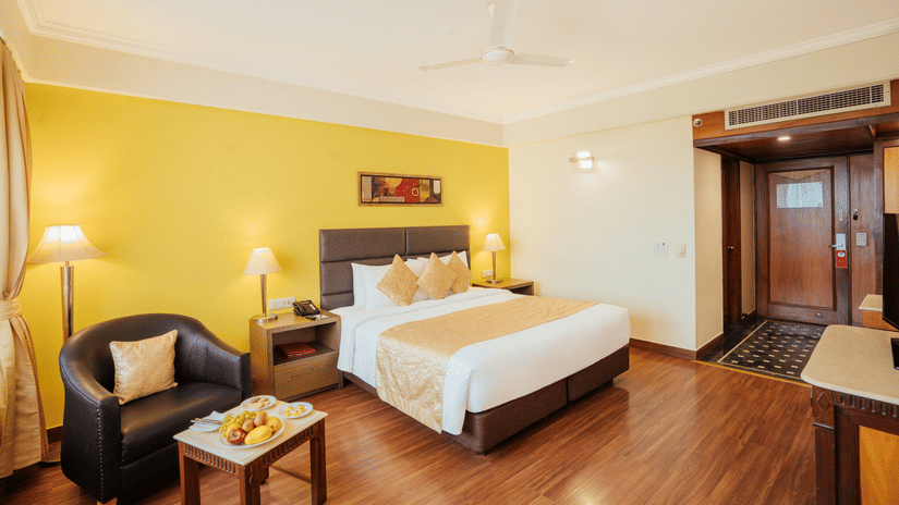 A modern bedroom with a neatly-made bed, yellow accent wall, leather armchair, coffee table with a fruit plate, and wooden flooring at The Piccadilly Lucknow.