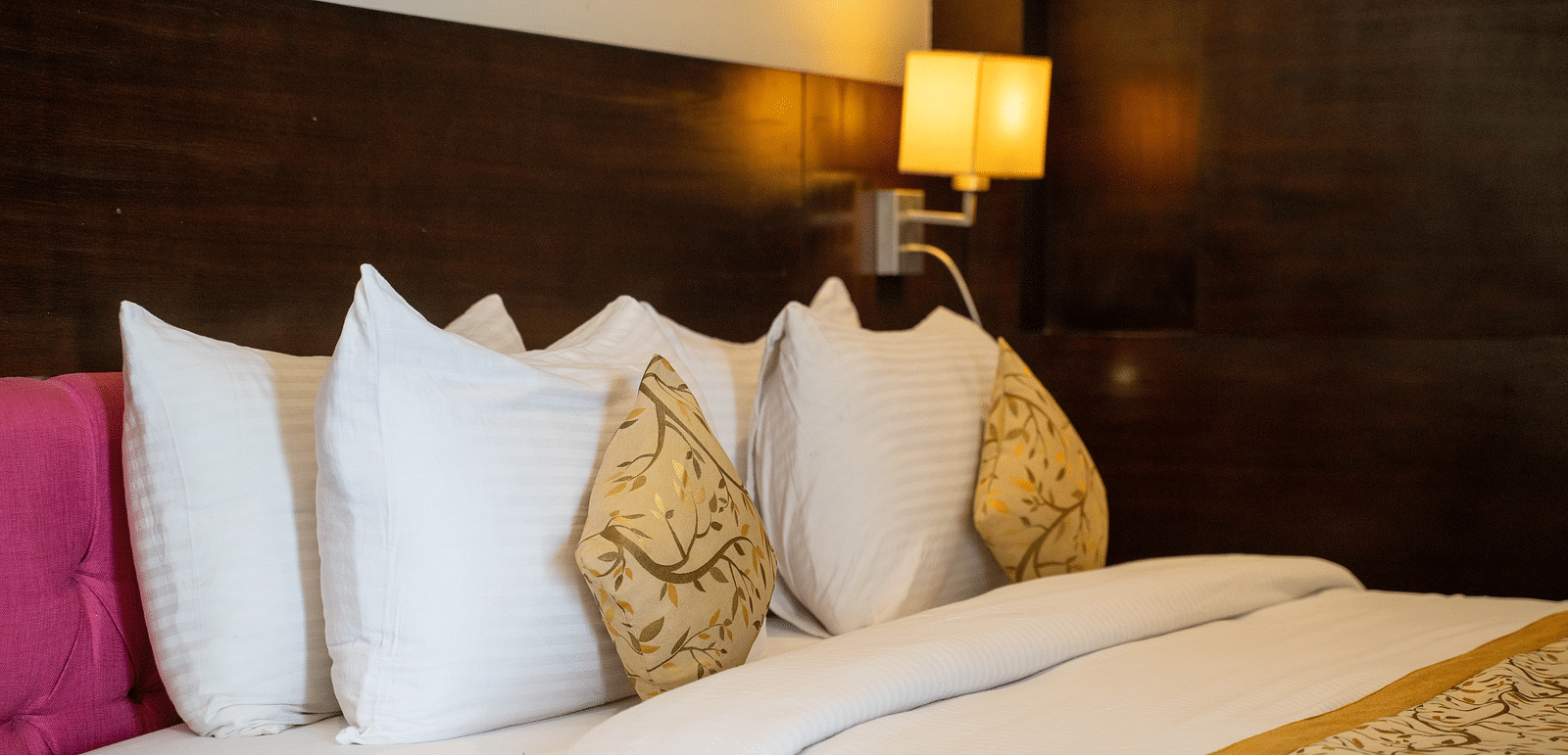 A neatly made bed with white linens and multiple pillows, including a patterned decorative pillow, is situated against a dark wood headboard, with a wall-mounted lamp emitting a warm light | Hotel Udai Median, Udaipur