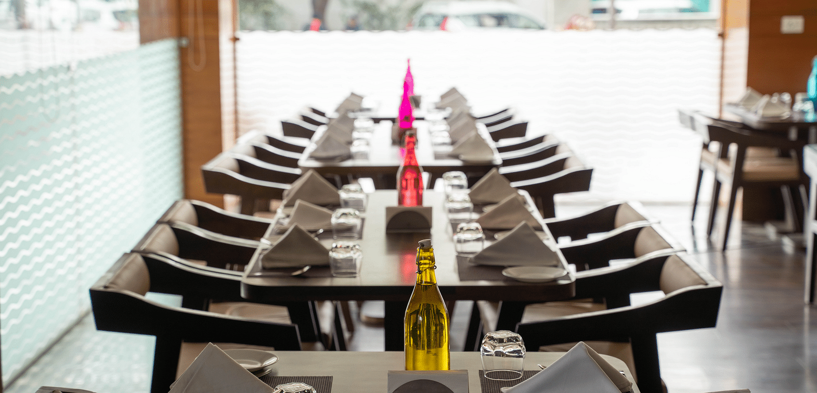 A long, set dining table in a restaurant features dark wooden chairs and place settings with folded napkins, glassware, and a decorative bottle | Hotel Udai Median, Udaipur