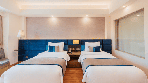 A twin room featuring 2 single beds, a wall panel headboard, bedside lamps, and warm ceiling lighting at The Piccadilly Lucknow.