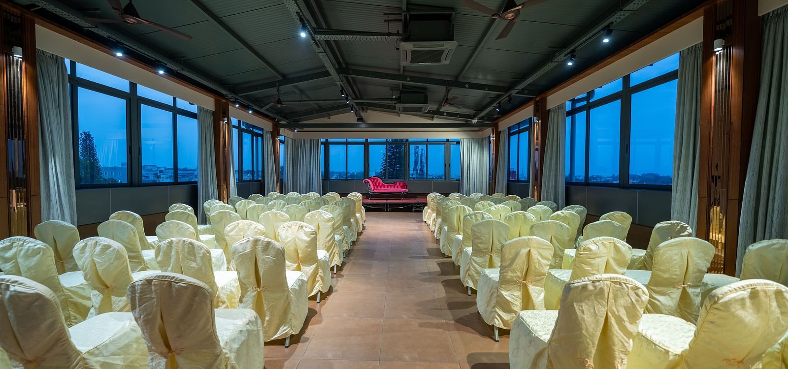 A view of the banquet hall at Hari Govinda's Express, Vijayanagar, with chairs arranged on either side of a central aisle leading to the stage, which features a cushioned sofa, while the curtains are drawn to the sides.