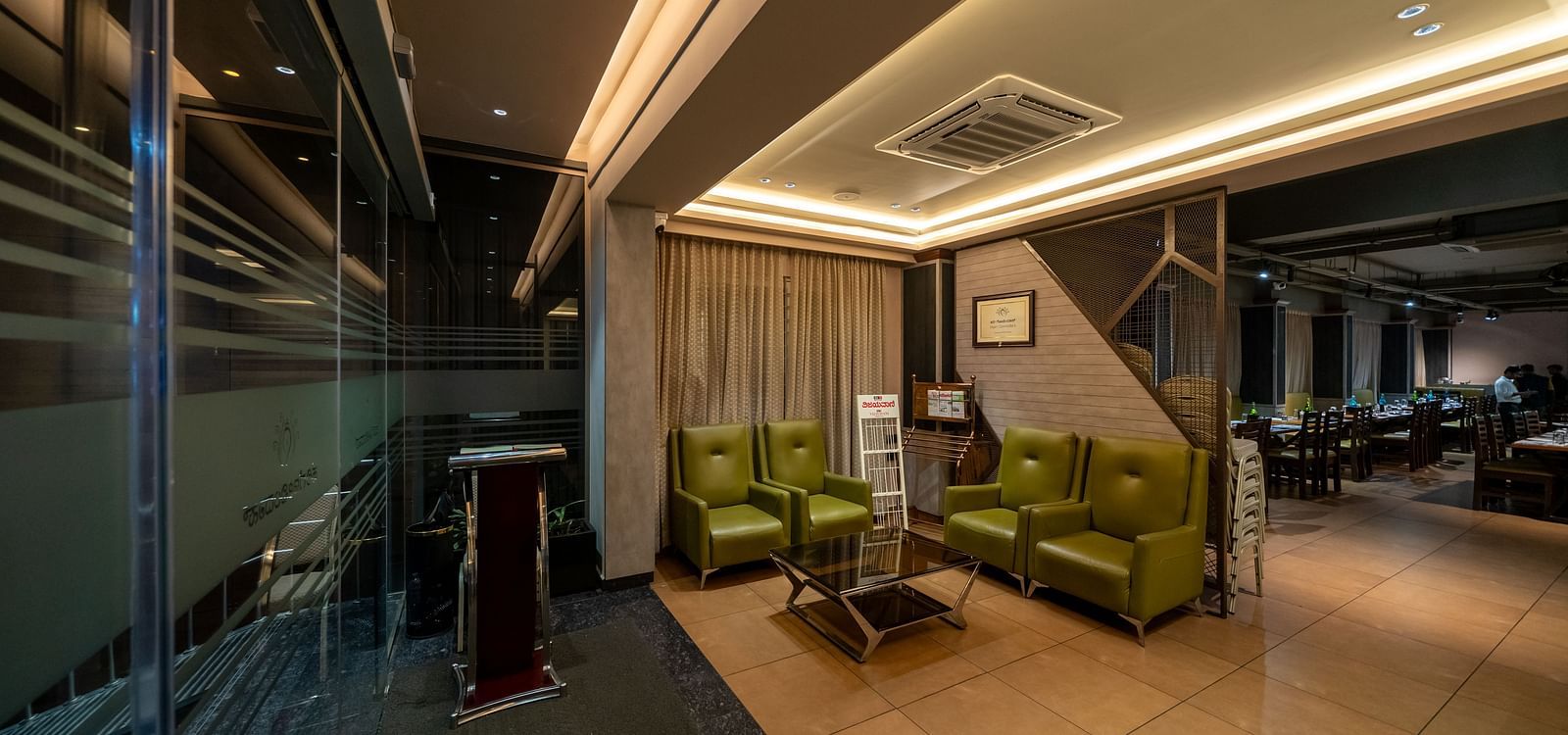 Lobby area at Hari Govinda's Express, Vijayanagar, features L-shaped cushioned sofas with a glass table in front, complemented by warm and welcoming lighting.