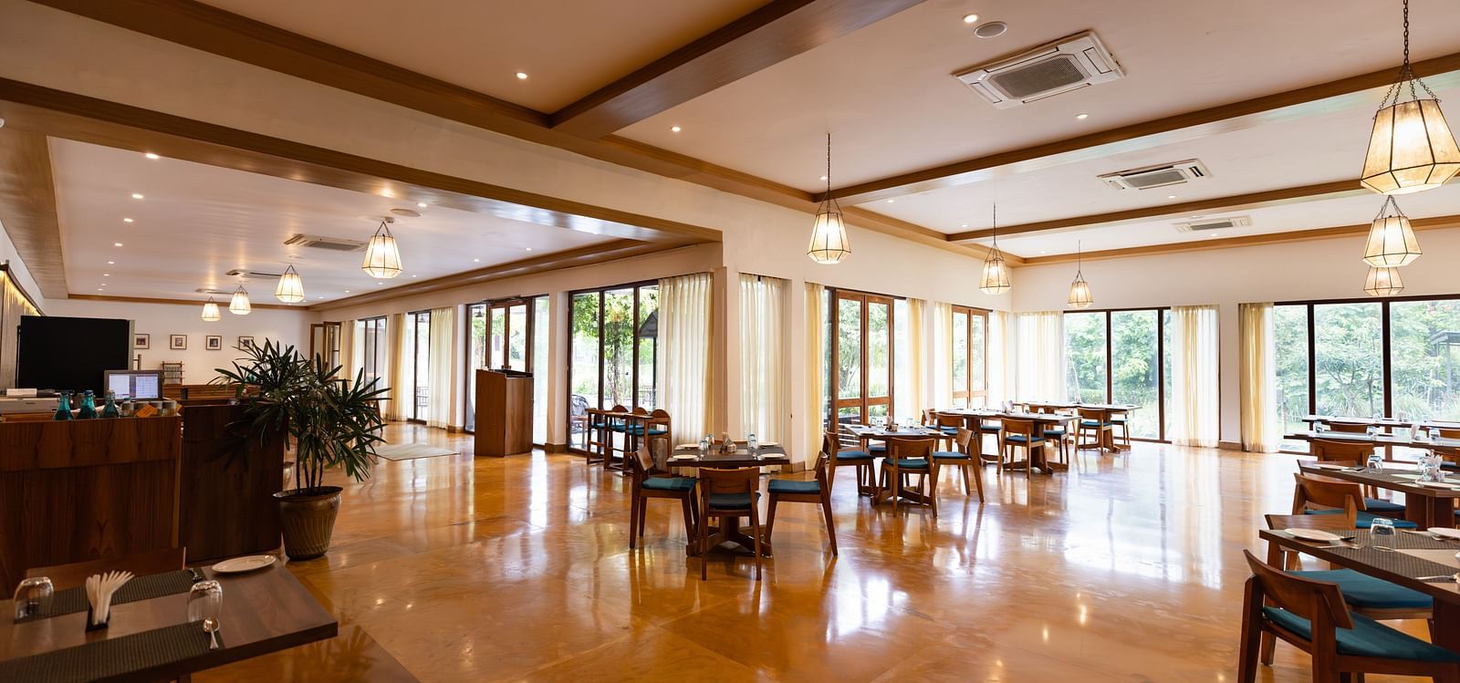 Seating arrangement in Azrak Restaurant at Saraca Resort & Spa Corbett with wooden flooring and wooden panelling on the ceiling alongside large French windows bringing in lots of light