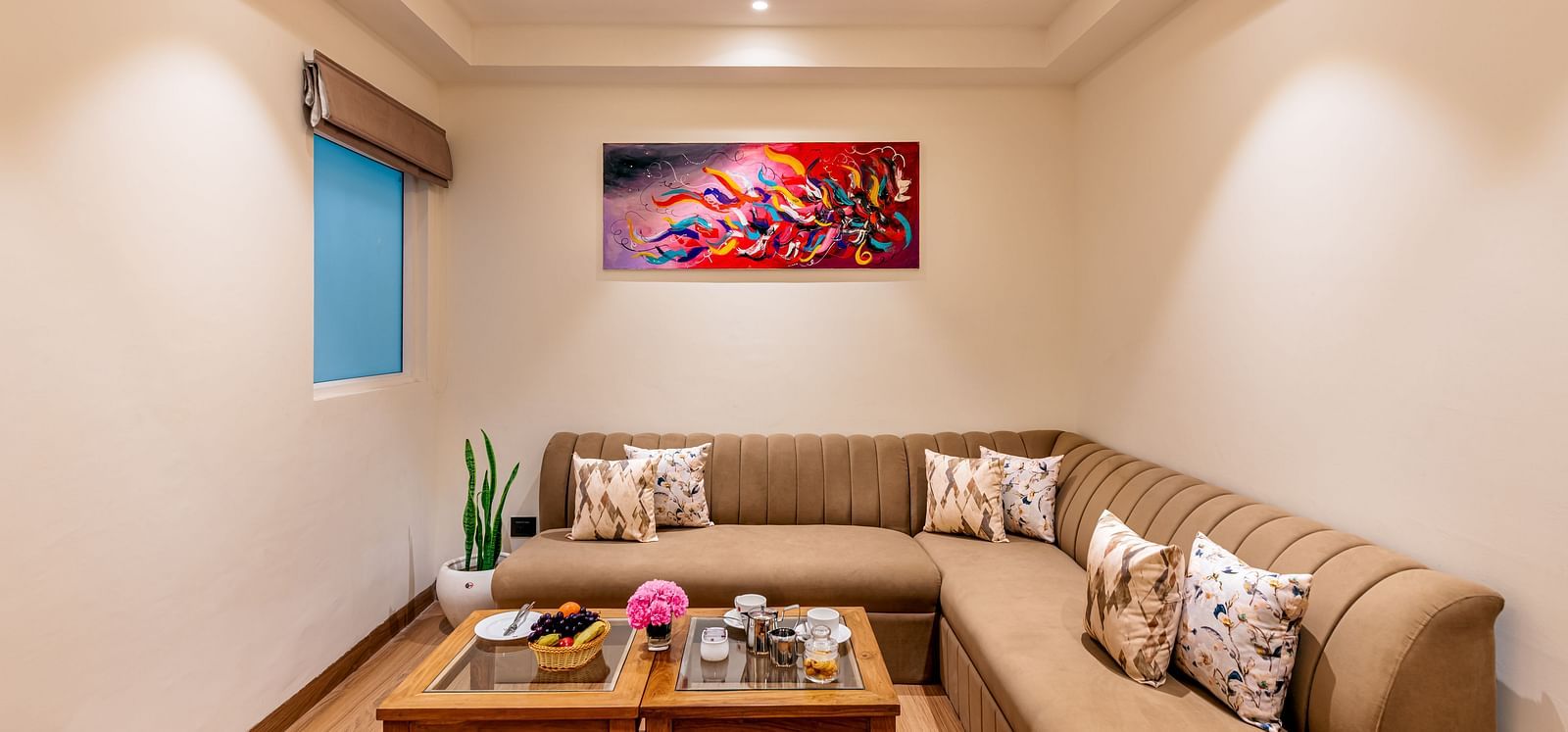 A living room with a sectional couch, a coffee table, and an abstract painting on the wall. It features a distinctive tray ceiling with lighting and a small window - Lords Eco Inn Dehradun.