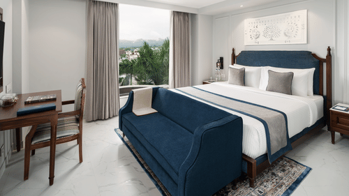 A room with a large bed with a blue bed runner, placed in front of a work desk, and a window offering a scenic view - MJ Sarovar Portico, Haridwar