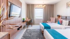 Premium room at Pride Elite Dwarka featuring twin beds, each with cushions, a flat TV screen, a work desk and chair, a window, and lights hanging from the ceilings.