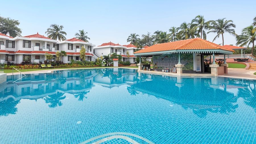 Wide swimming pool reflecting the clear sky, lined with villas and palm trees.