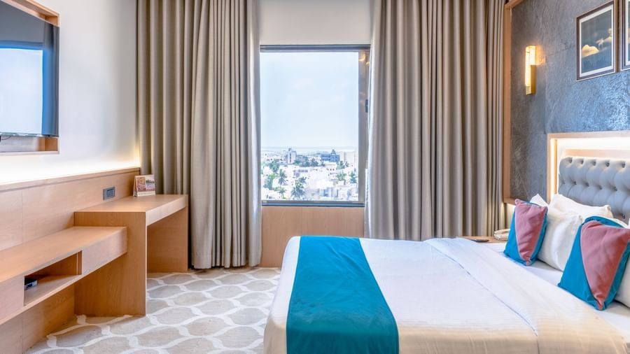 Deluxe Room at Pride Comfort Dwarka featuring  a double size bed, a cabinet table, a flat TV screen, and a window with curtains.