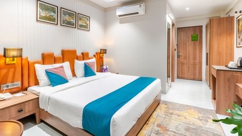 Deluxe room at Pride Elite Dwarka showcasing double size bed, an AC, a bedside table with lamp, a cabinet table, and a wardrobe.