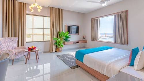 Suite at Pride Elite Dwarka featuring a bed, two windows, flat TV screen, a sofa chair with centre table, a flat TV screen, lights hanging from the ceiling, and a flower pot.