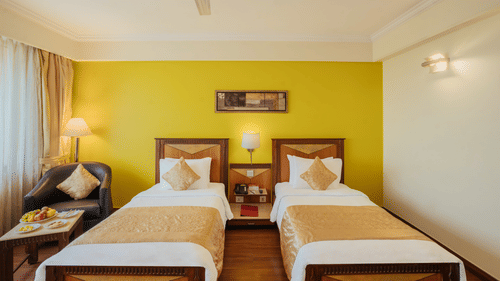 A bedroom featuring twin beds, a nightstand between the beds, and a leather sofa next to the window at The Piccadily Lucknow.