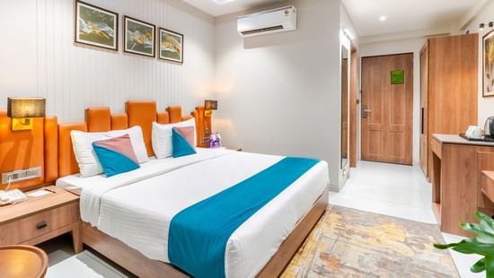 Deluxe room at Pride Comfort Dwarka showcasing double size bed, an AC, a bedside table with lamp, a cabinet table, and a wardrobe.