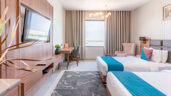 Premium room at Pride Comfort Dwarka featuring twin beds, each with cushions, a flat TV screen, a work desk and chair, a window, and lights hanging from the ceilings.