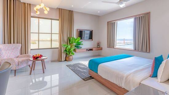 Suite at Pride Comfort Dwarka featuring a bed, two windows, flat TV screen, a sofa chair with centre table, a flat TV screen, lights hanging from the ceiling, and a flower pot.