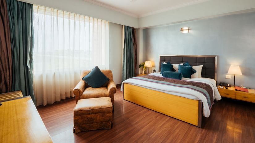 A room at The Piccadily Lucknow hotel featuring a double size bed, a sofa chair, bedside table with lamps on either side, and a large window with curtains.