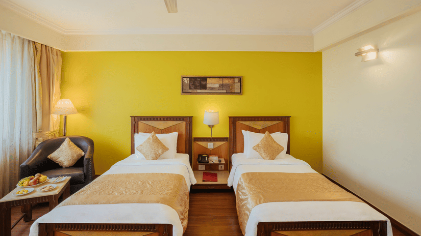 A bedroom featuring twin beds, a nightstand between the beds, and a leather sofa next to the window at The Piccadily Lucknow.