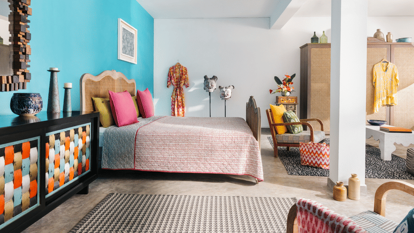 A spacious, brightly lit bedroom at Owl and the Pussycat Hotel in Galle featuring a bed with textured linens and vibrant pillows, an armchair with a woven basket, and a decorative sideboard showcasing a geometric pattern.