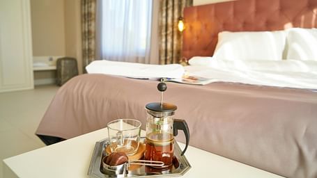 Well-lit hotel bedroom with king-size bed and a coffee tray featuring a French press, glass mug, and chocolate on a small table near the window.