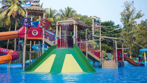 A colourful pirate-themed water playground with slides, splash features, and a skull-shaped structure at a water park, surrounded by palm trees.
