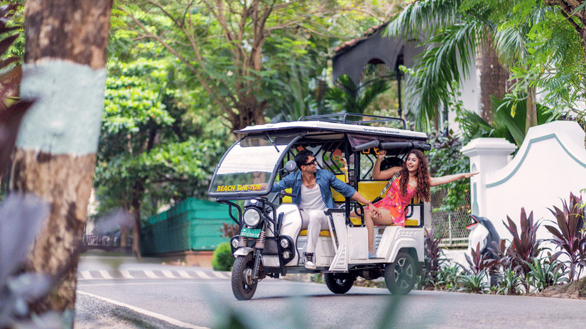 A couple on a colourful tuk-tuk parked under lush trees at a tropical resort entrance | Heritage Village Resorts & Spa, Goa