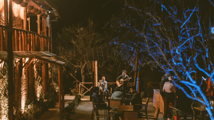  A lively outdoor gathering with people seated around a musician playing a guitar during a live music session at Boca Mario inside The Manor Luxury Hotel, Shimla.