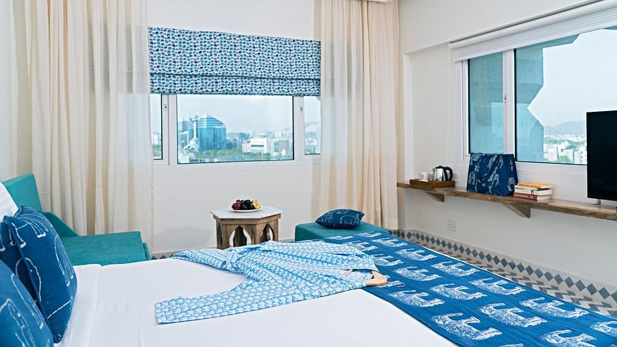 A bright hotel room with a king-size bed featuring blue patterned bedding, a turquoise armchair, and city views from the windows at Clarks Amer, Jaipur.