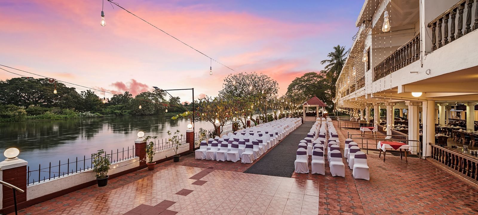 An image of the outdoor event space with seating arrangement near a lake with bright decorative lighting - GReaT Trails River View Resort Thanjavur By GRT Hotels