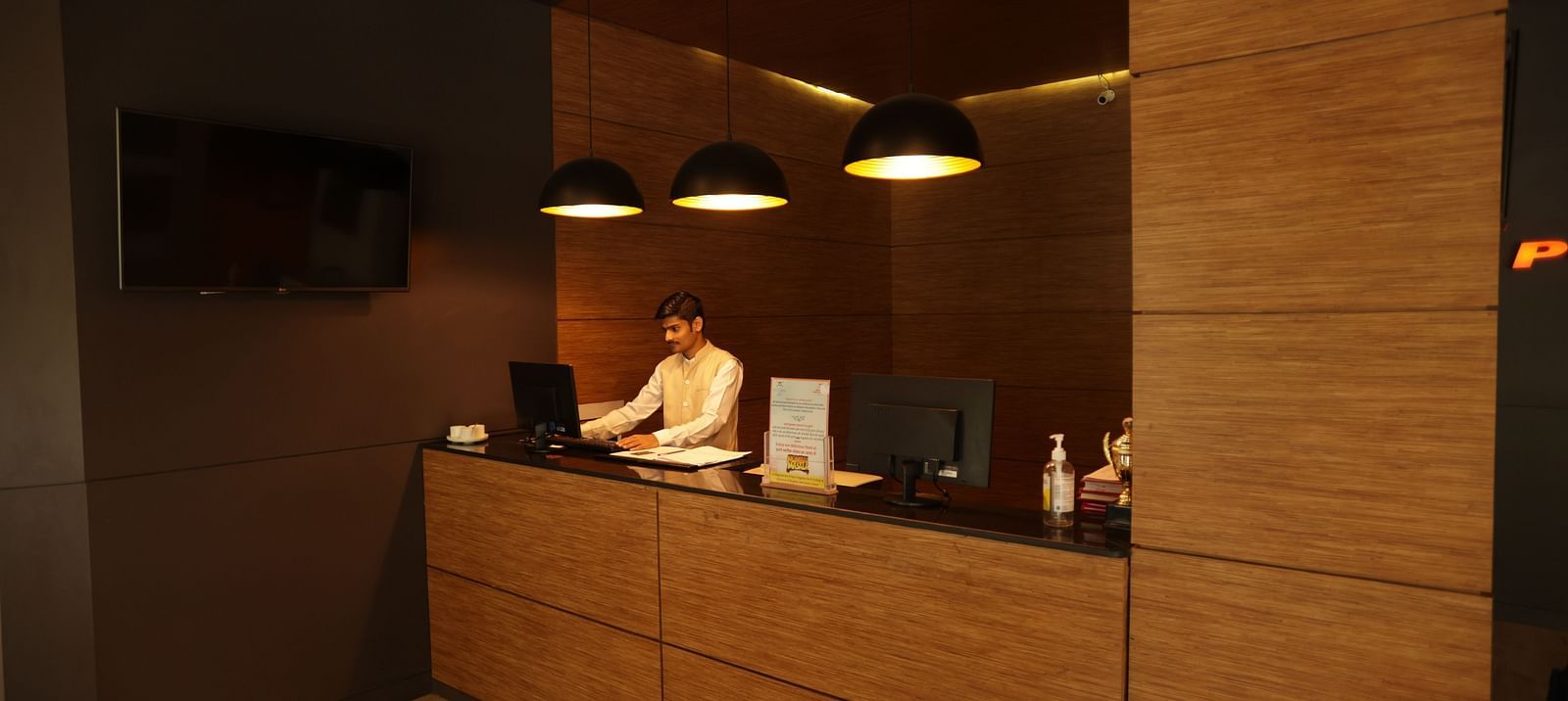 A person is front of the laptop at the front desk of the Max Hotel Jabalpur, featuring a wood-paneled reception area and warm lighting. 