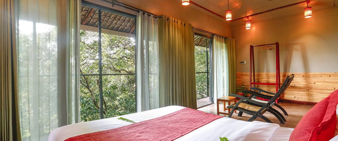 A bed placed in a room with tall glass windows and ambient lighting | Vythiri Resort, Wayanad