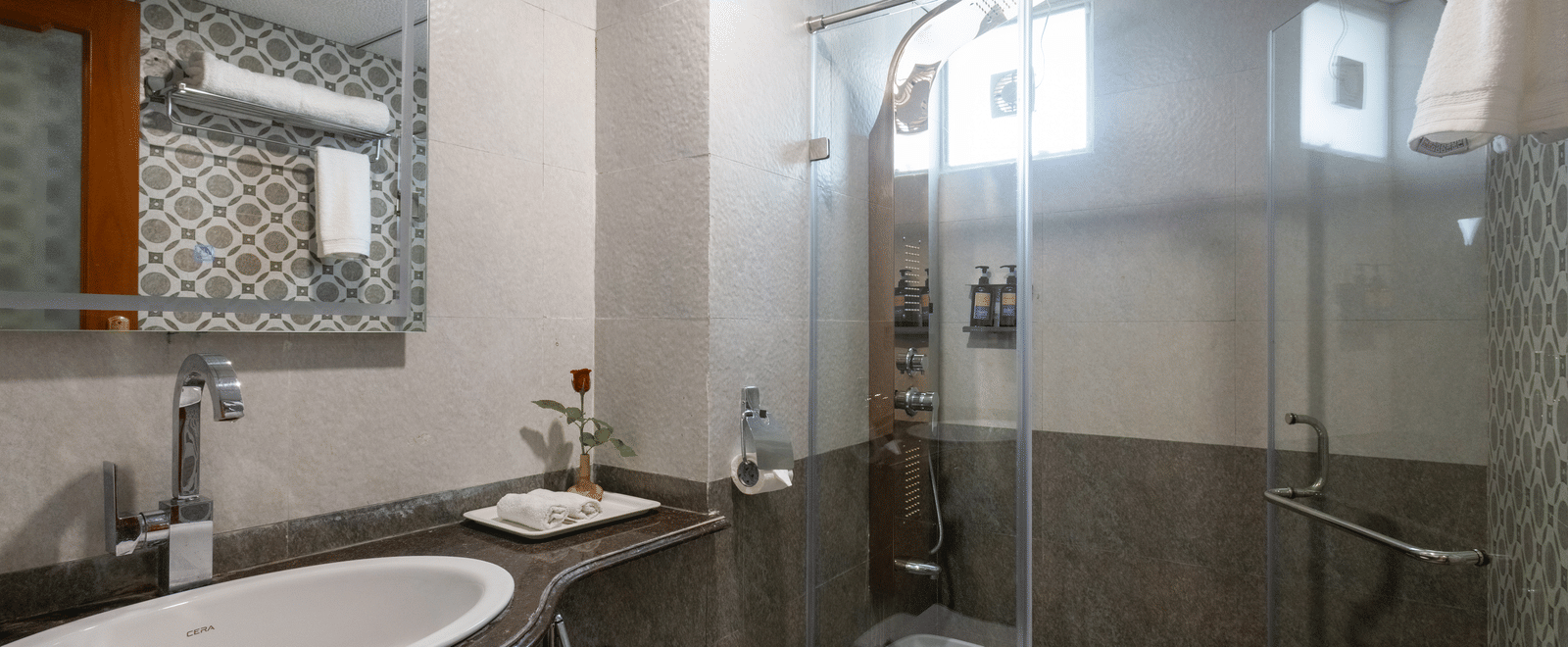 Stylish hotel bathroom showing a glass shower enclosure, a white basin, and an ornate framed mirror on the wall at Pravasa Stay, Vadodara.