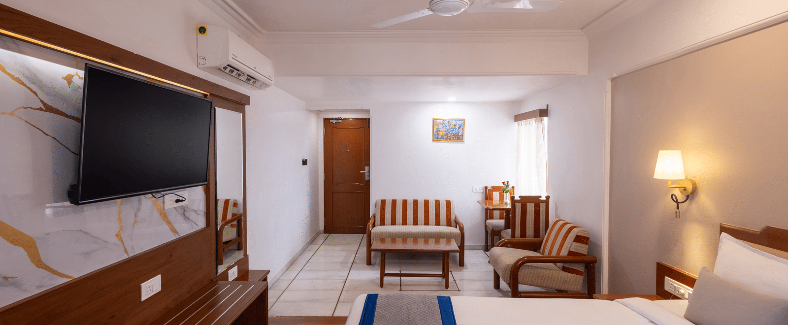 A warm, spacious hotel room with a wooden floor, a large bed, and a seating area visible near the window at Pravasa Stay, Vadodara.