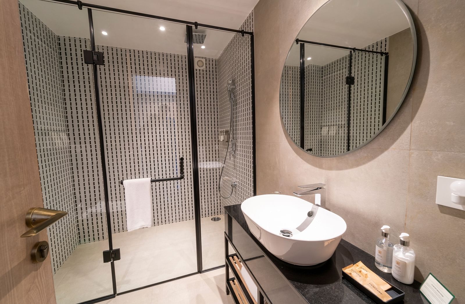 Side view of the washroom showing a spacious washbasin under a round mirror, with a glass partition enclosing the showering space at Nivasana Manali.