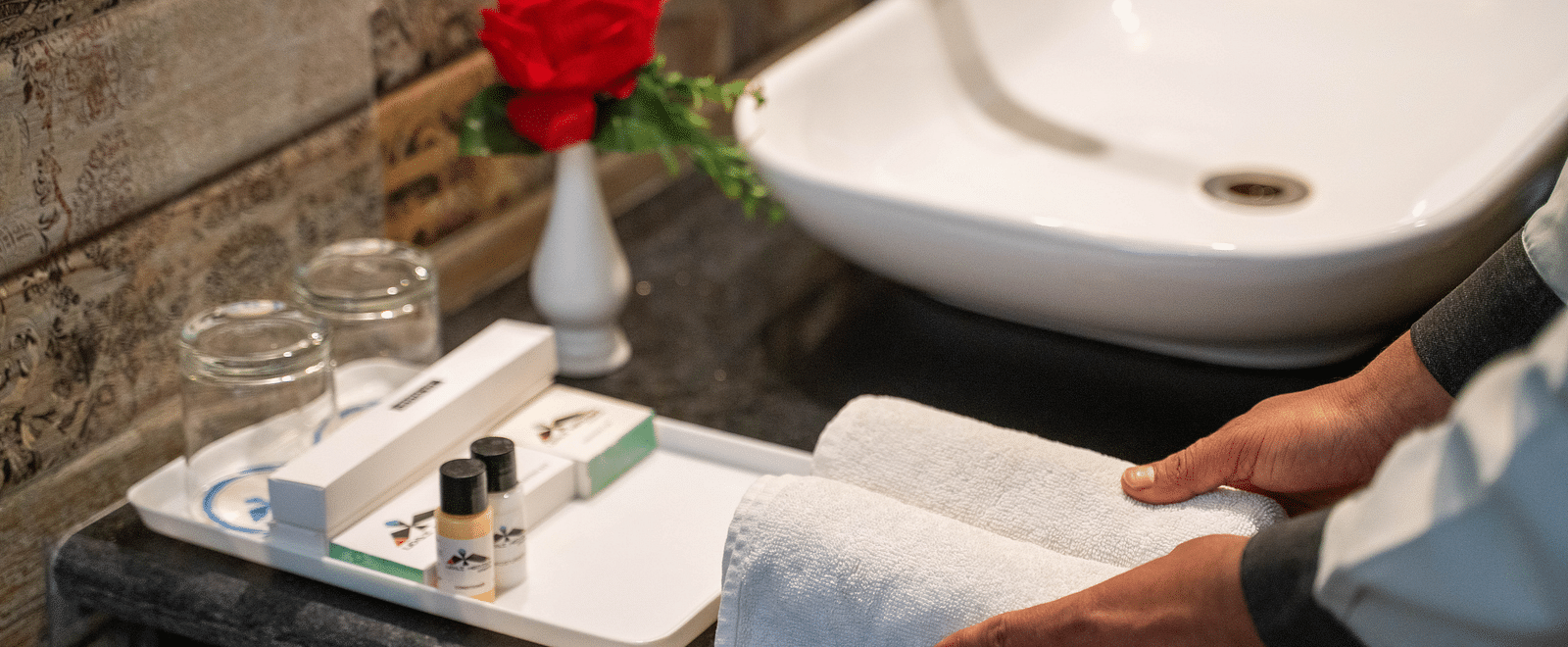 A staff member preparing a bathroom counter with fresh towels, toiletries, and a flower vase - Hotel Udai Median, Udaipur.