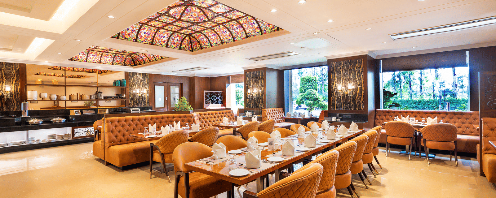 Long view of the spacious restaurant with multiple dining tables, comfortable seating, and a view of the outside through windows at Hotel Hukam's Lalit Mahal.