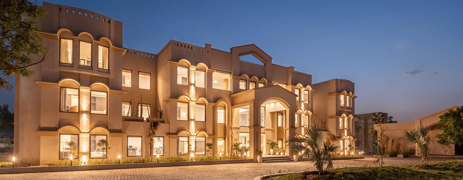 An image of a well lit facade with a huge concrete lawn and a few trees - Brij Eternity, Vrindavan