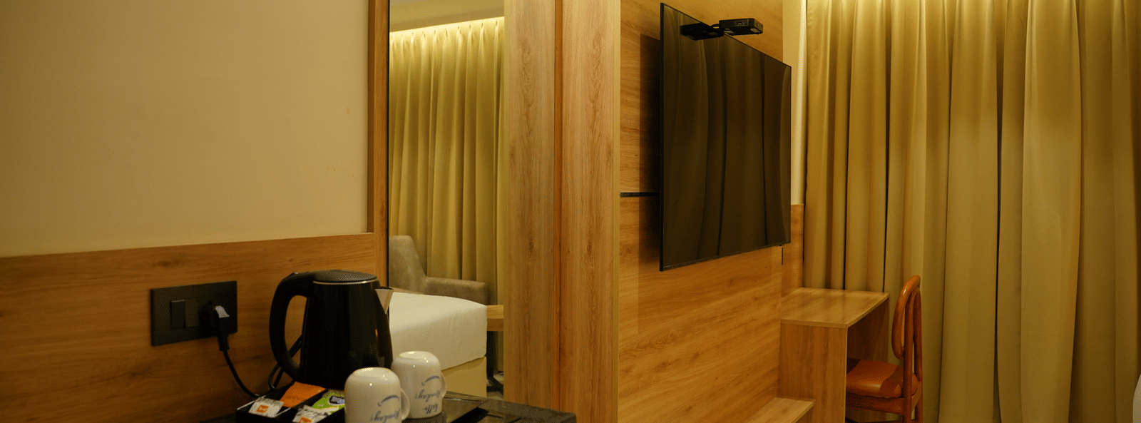  Modern guest room setup at Monday Premium, Madhapur, showing a TV on a wood-panelled wall and tea-making facilities nearby.