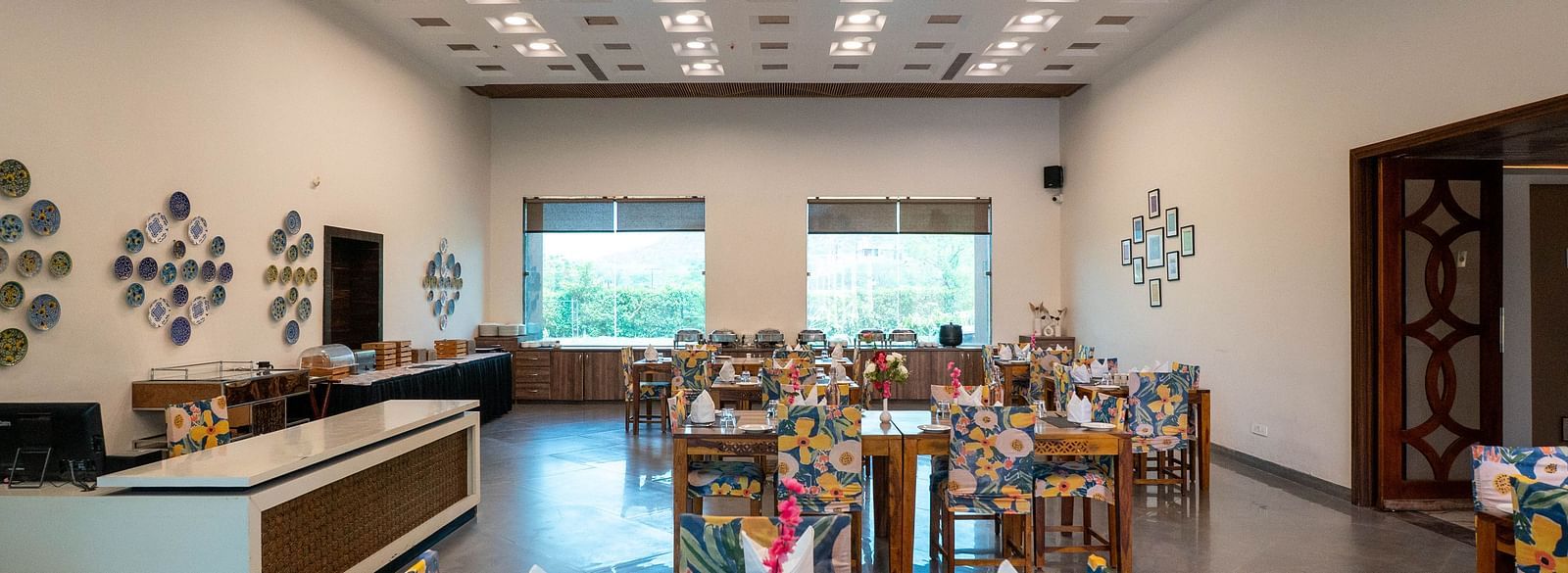 Bay Leaf Restaurant at Bamboo Saa Mulberry Resort in Pushkar with neatly lined tables, a help desk and two large glass windows.