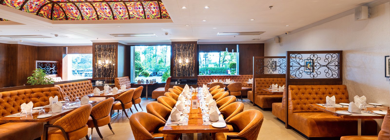 Restaurant dining hall featuring orange booths, wooden tables set with orange cloths, and a decorative ceiling panel at Hotel Hukam's Lalit Mahal.