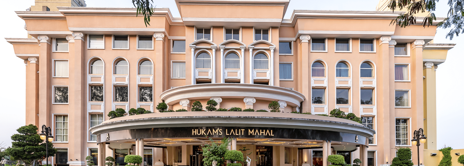 Large, modern, multi-storey building in a peachy-pink colour with a curved canopy entrance and formal landscaping at Hotel Hukam's Lalit Mahal.