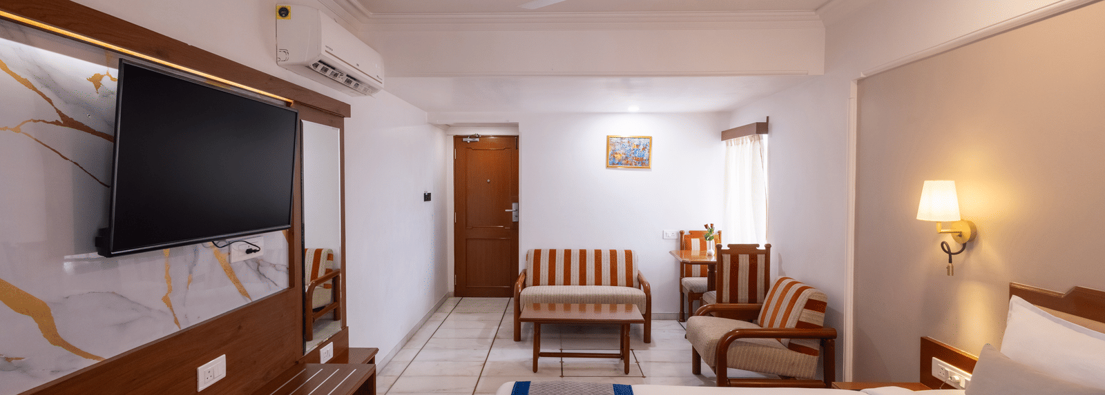 A warm, spacious hotel room with a wooden floor, a large bed, and a seating area visible near the window at Pravasa Stay, Vadodara.