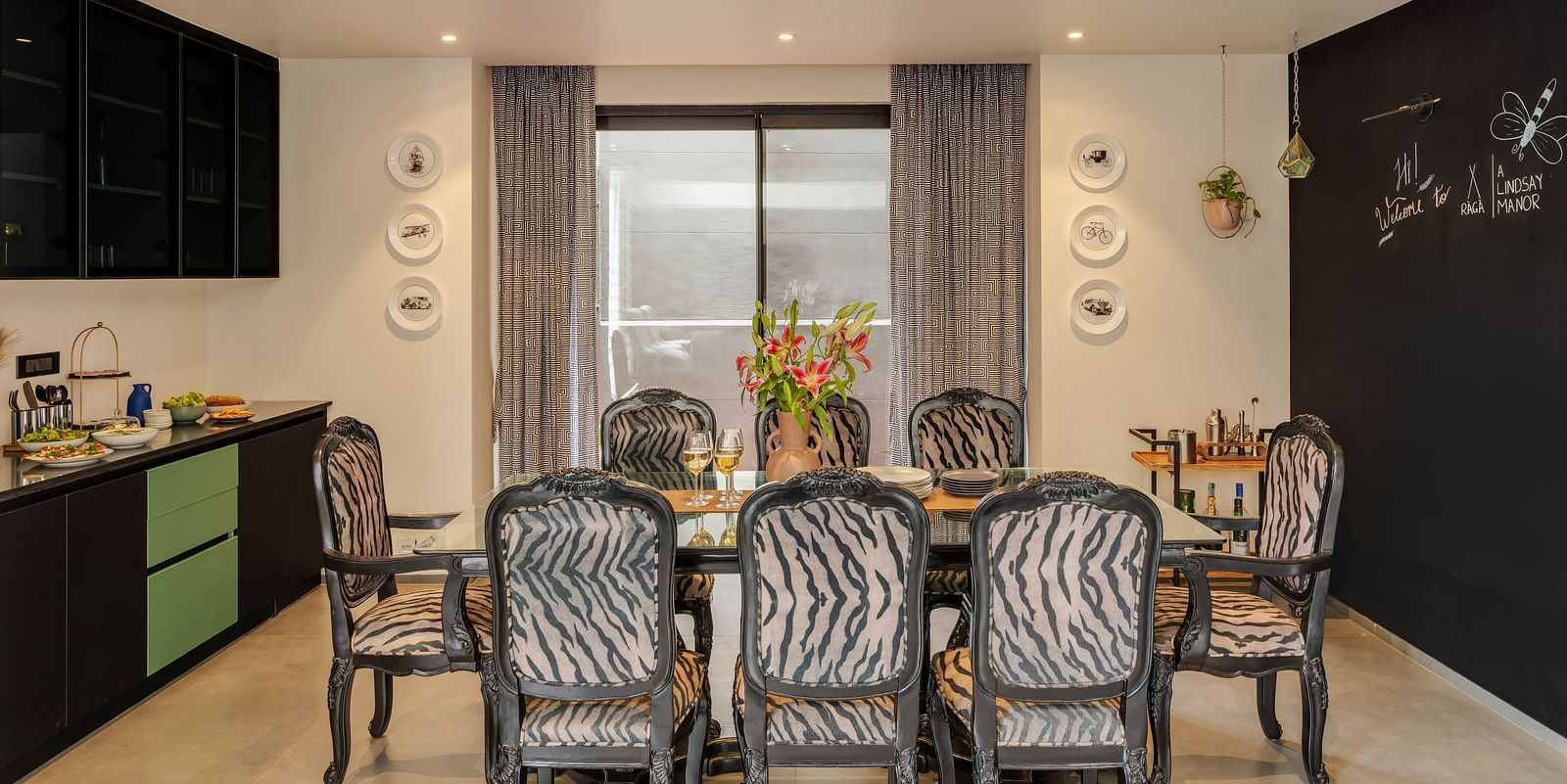 A dining area at Lindsay Manor featuring a table with chairs, a kitchenette beside, and a window behind the table.
