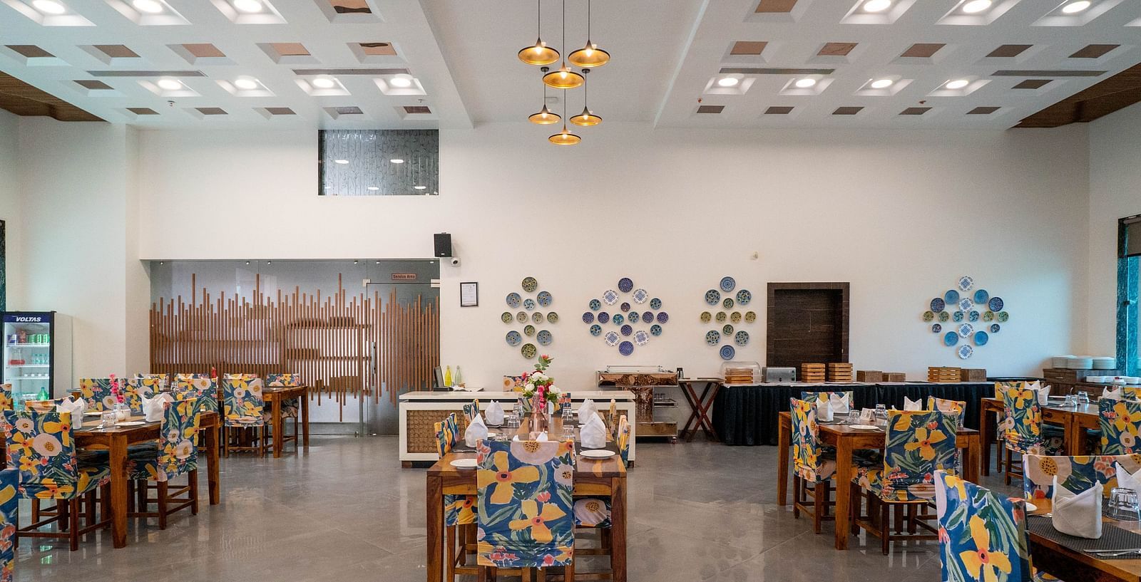 The Bay Leaf Restaurant at Bamboo Saa Mulberry Resort in Pushkar featuring  neatly arranged rows of dining tables with overhead lighting and a beverage vending machine in the corner.